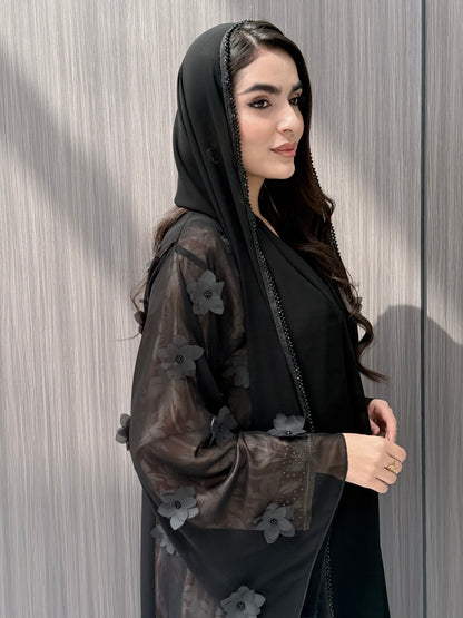 Woman wearing an open-front sheer black abaya featuring elegant 3D floral appliqués. Complete with a matching beaded sheila, perfect for weddings, Eid, and special occasions standing on a wooden floor.