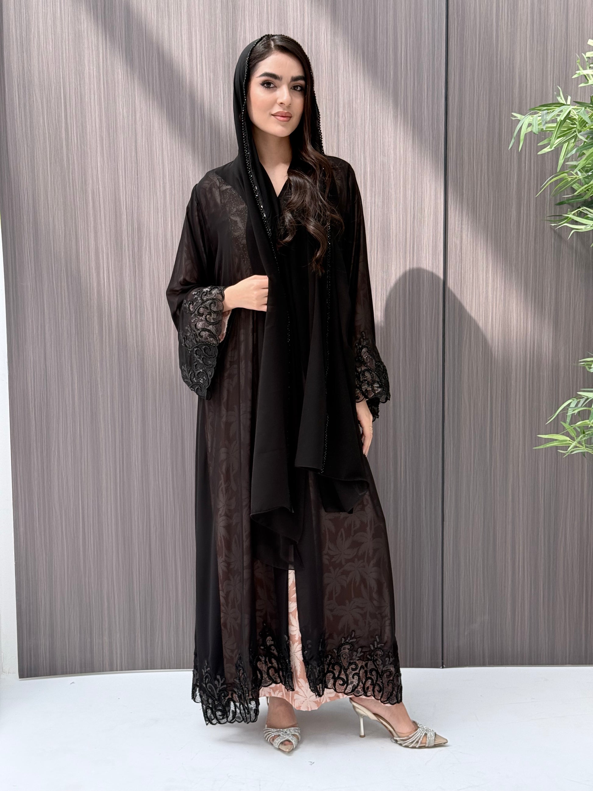 Woman wearing an open-front sheer black abaya with a matching beaded sheila on a gray background