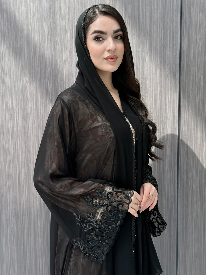Woman wearing an open-front sheer black abaya with a matching beaded sheila on a gray background