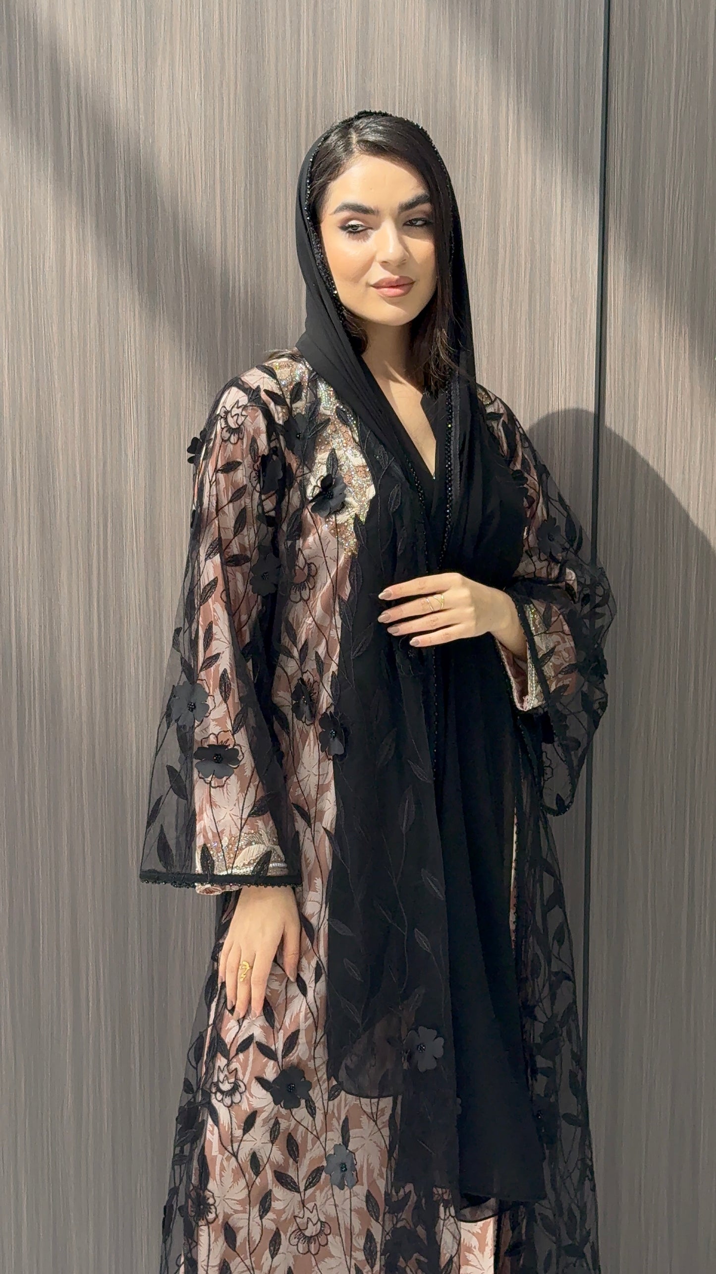 Woman wearing a Black Sheer Floral Appliqué Open Front Abaya with a mathcing sheila against a wooden panel background