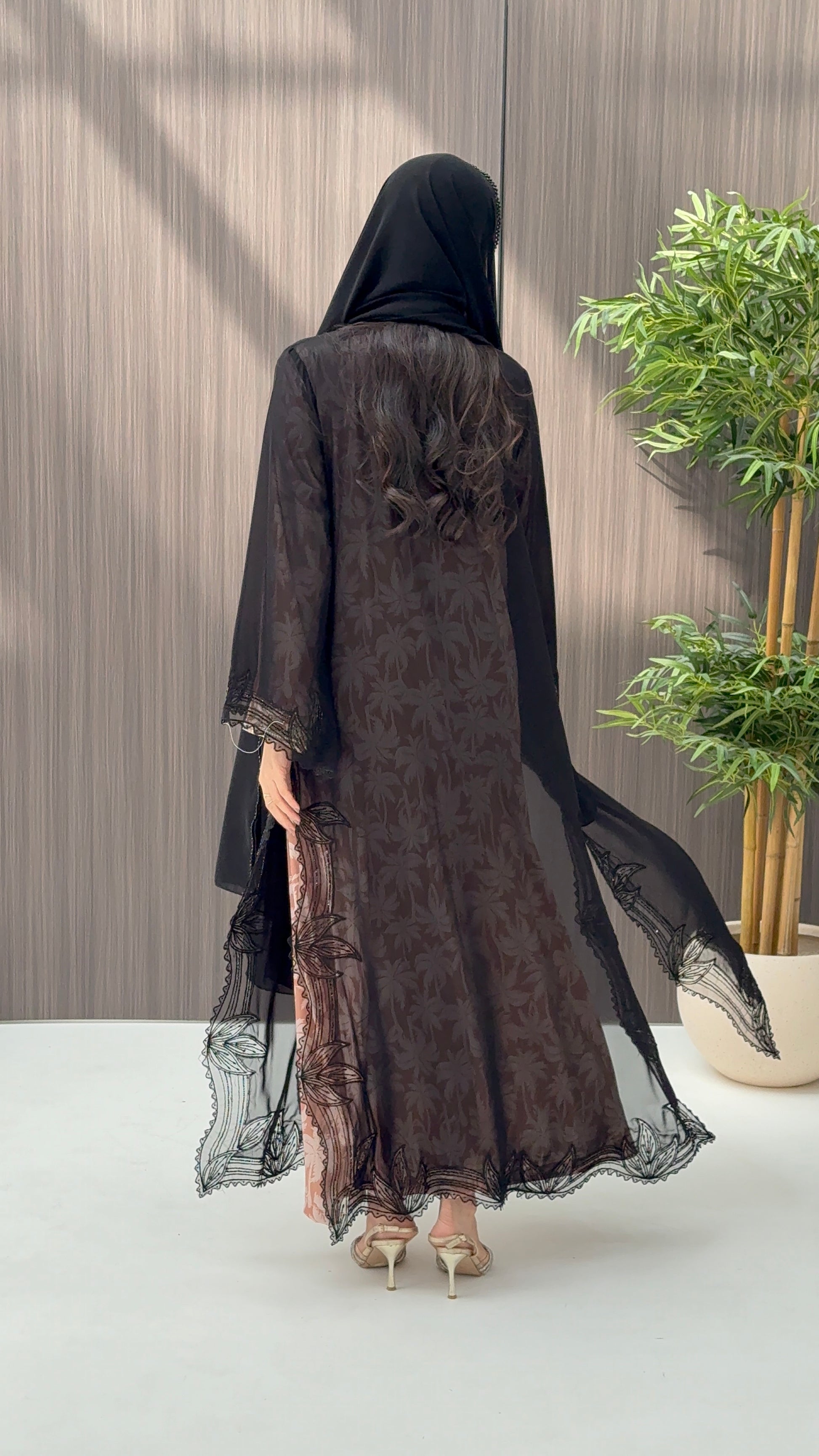 A woman wearing a luxury open-front abaya crafted from premium Nida fabric with a sheer botanical overlay and elegant beaded leaf lace appliqués. Includes a matching beaded sheila in a minimal indoor setting.