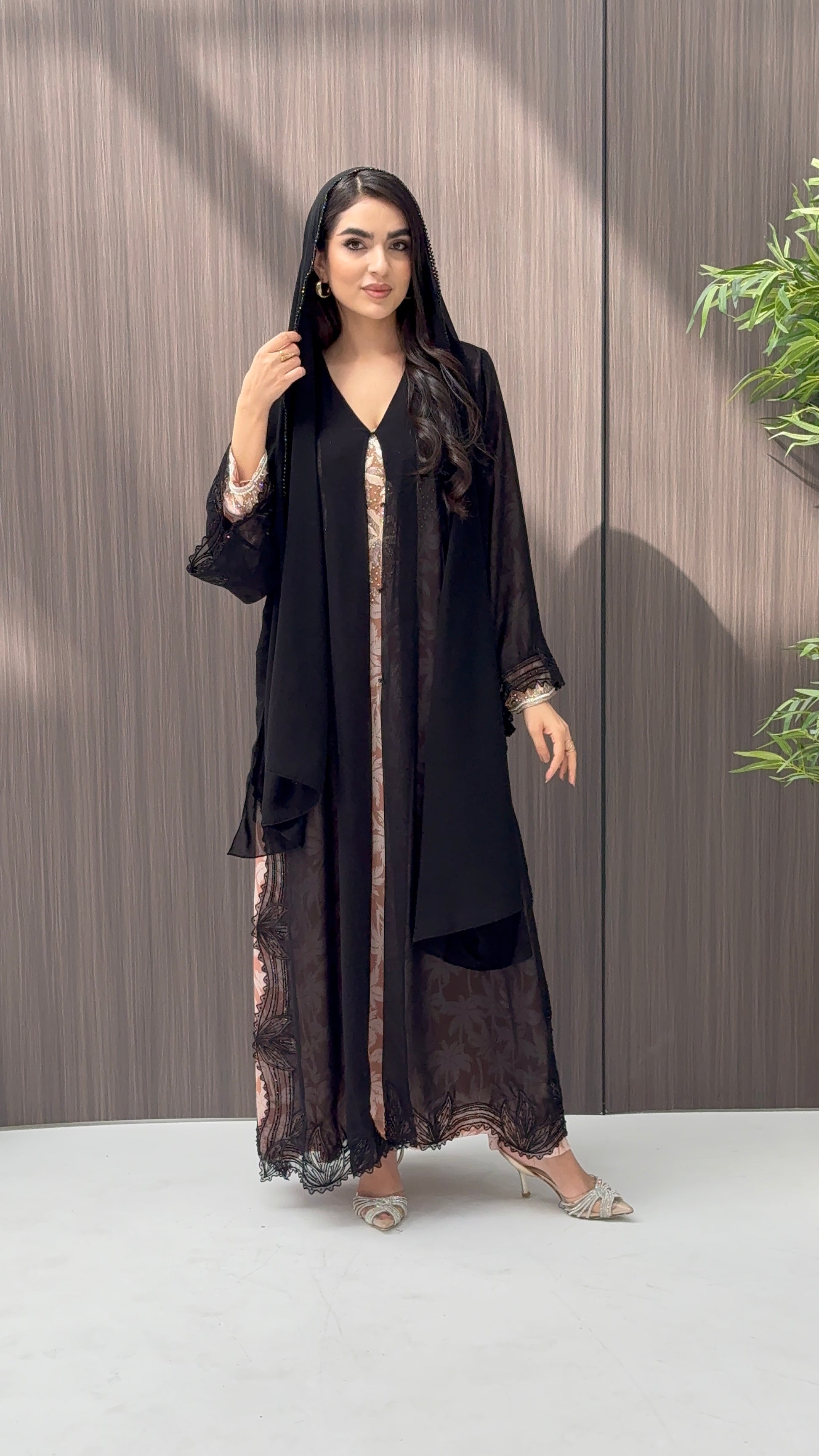 A woman wearing a luxury open-front abaya crafted from premium Nida fabric with a sheer botanical overlay and elegant beaded leaf lace appliqués. Includes a matching beaded sheila in a minimal indoor setting.
