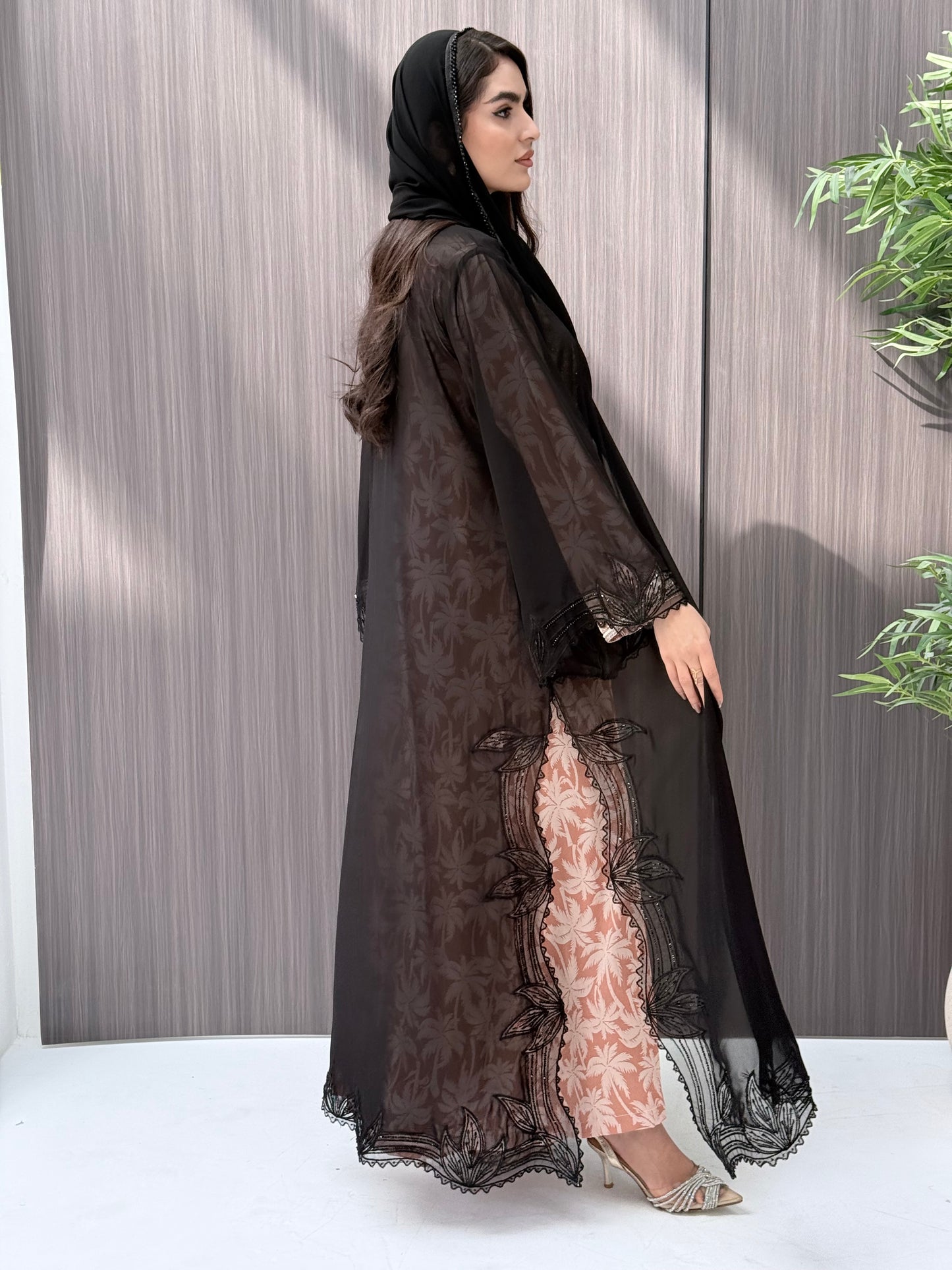 A woman wearing a luxury open-front abaya crafted from premium Nida fabric with a sheer botanical overlay and elegant beaded leaf lace appliqués. Includes a matching beaded sheila in a minimal indoor setting.