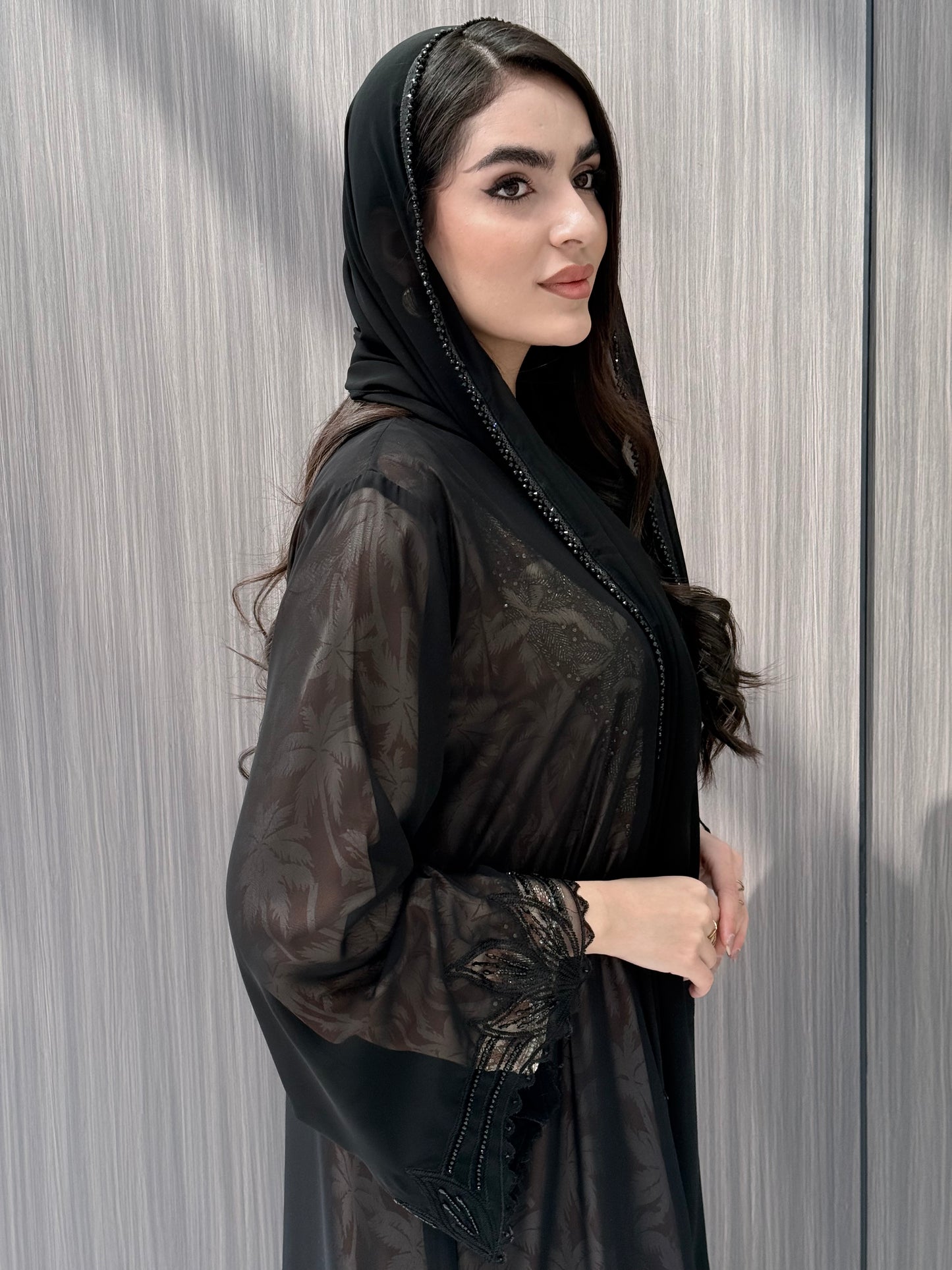 close up of a woman wearing a luxury open-front abaya crafted from premium Nida fabric with a sheer botanical overlay and elegant beaded leaf lace appliqués. Includes a matching beaded sheila in a minimal indoor setting.