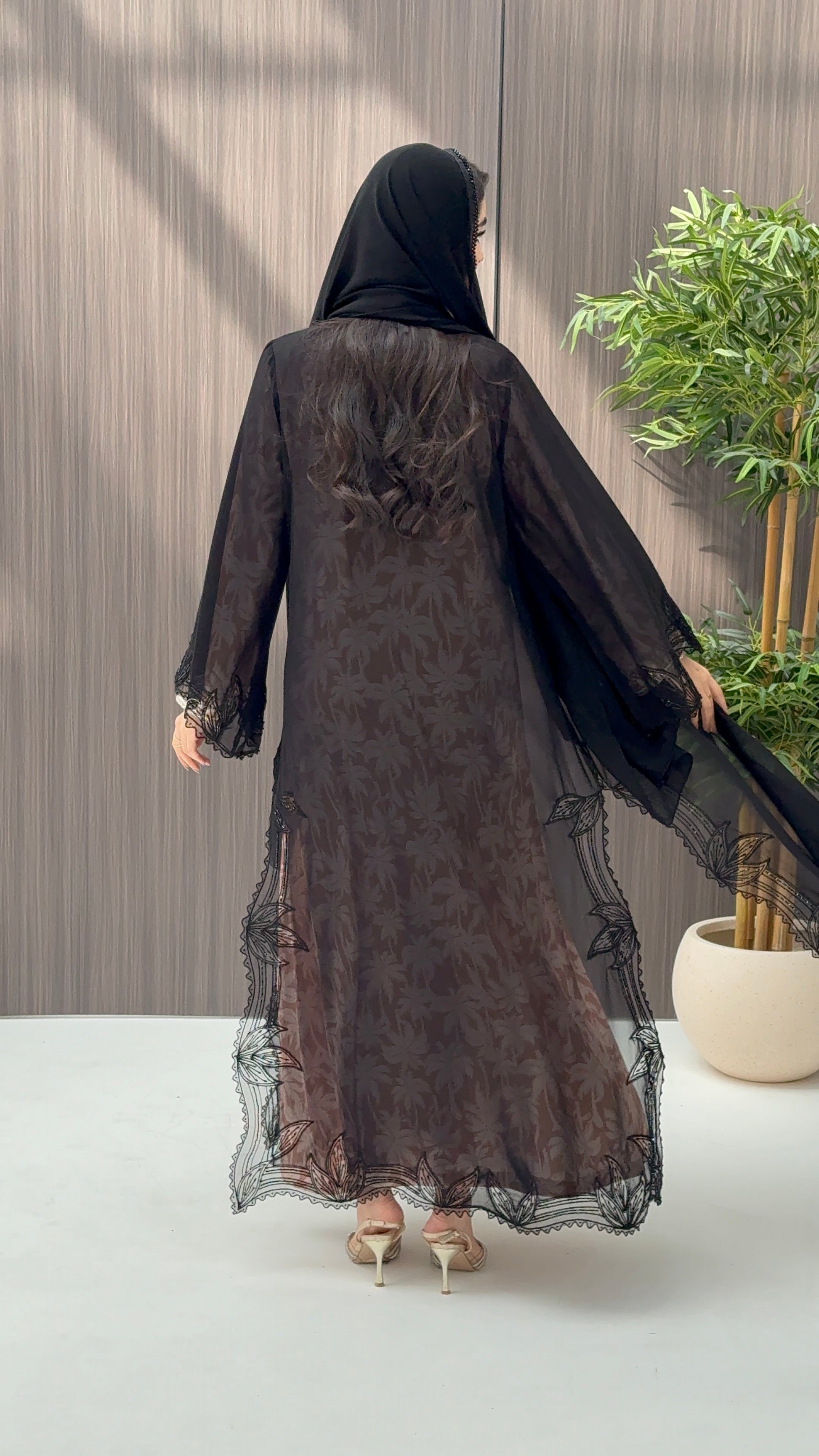A woman wearing a luxury open-front abaya crafted from premium Nida fabric with a sheer botanical overlay and elegant beaded leaf lace appliqués. Includes a matching beaded sheila in a minimal indoor setting.