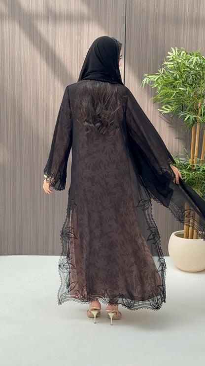 A woman wearing a luxury open-front abaya crafted from premium Nida fabric with a sheer botanical overlay and elegant beaded leaf lace appliqués. Includes a matching beaded sheila in a minimal indoor setting.