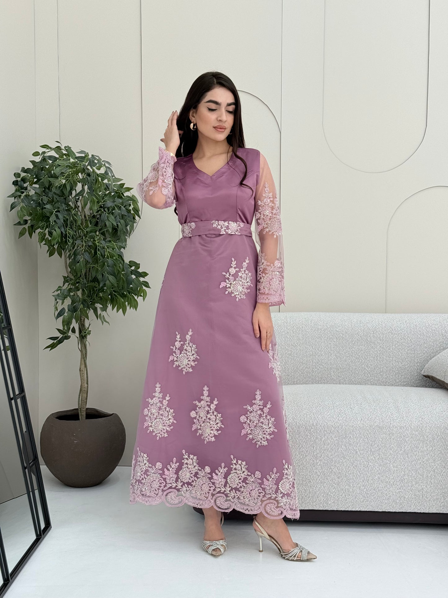 Woman in an elegant dusty purple modest jalabiya designed with a flattering V-neck, floral embroidered sheer sleeves, and a beautifully structured matching waist belt. standing against a white wall.