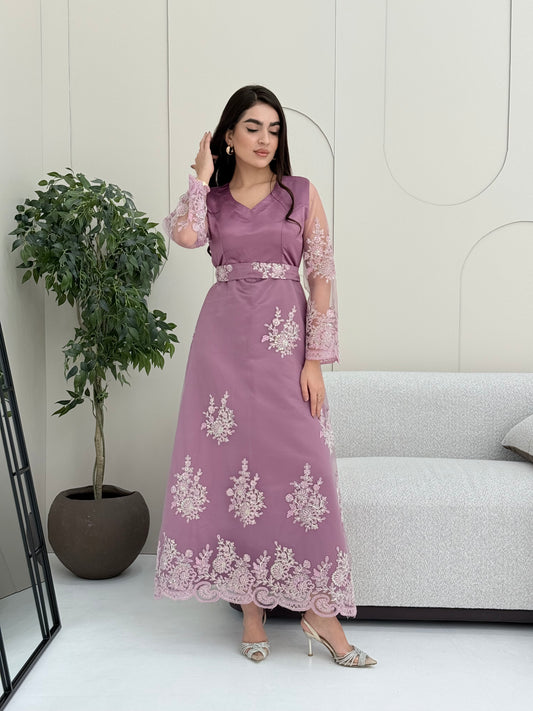 Woman in an elegant dusty purple modest jalabiya designed with a flattering V-neck, floral embroidered sheer sleeves, and a beautifully structured matching waist belt. standing against a white wall.