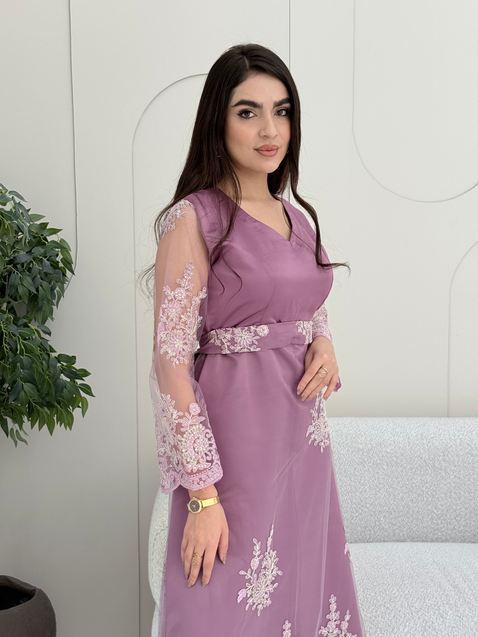 Woman in an elegant dusty purple modest jalabiya designed with a flattering V-neck, floral embroidered sheer sleeves, and a beautifully structured matching waist belt. standing against a white wall.