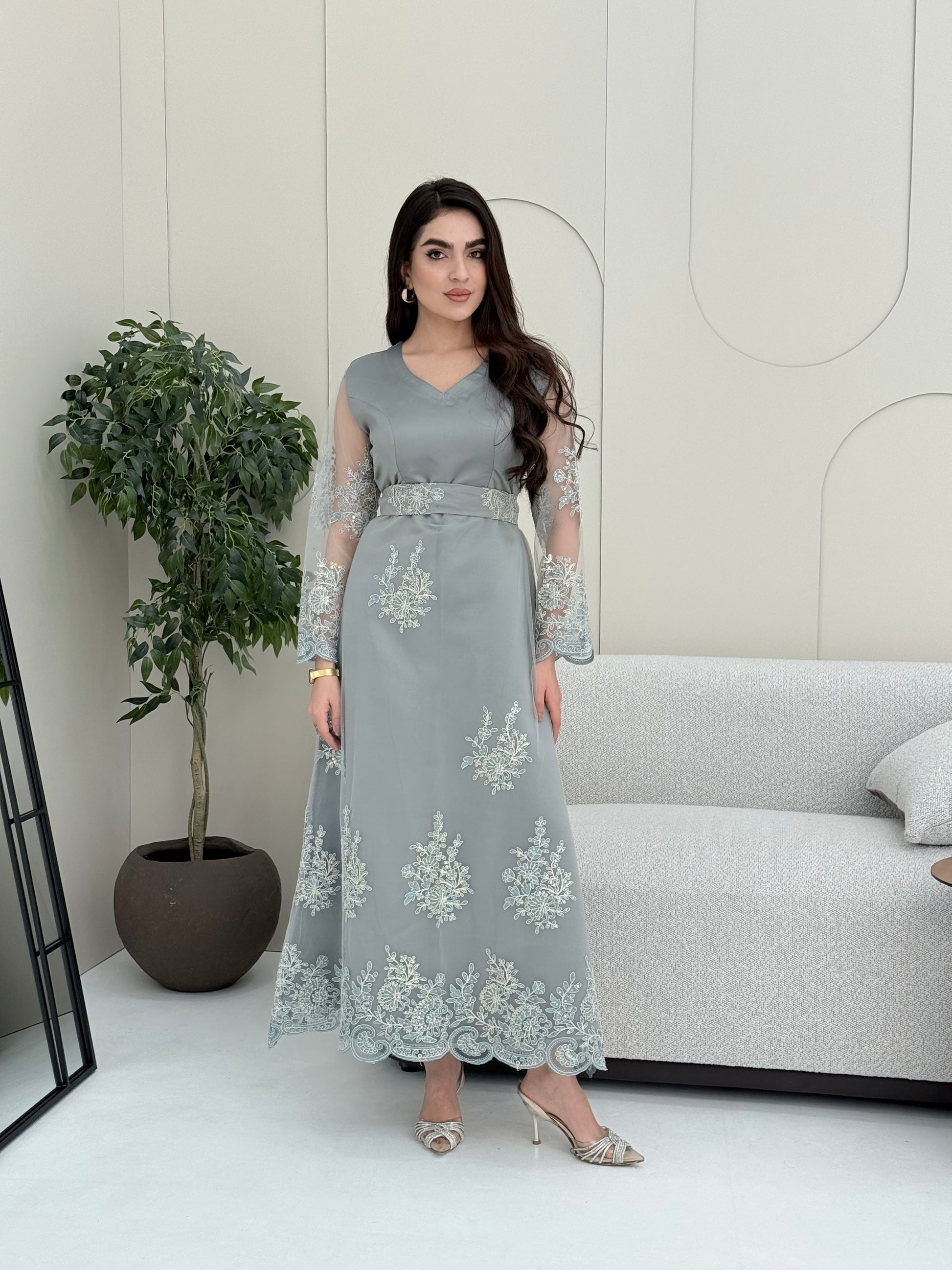 Woman wearing a light grey luxury jalabiya featuring delicate floral lace sheer sleeves, a tailored V-neck, and an elegantly embroidered matching waist belt, standing against a white wall with a plant in the foreground.