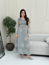 Woman wearing a light grey luxury jalabiya featuring delicate floral lace sheer sleeves, a tailored V-neck, and an elegantly embroidered matching waist belt, standing against a white wall with a plant in the foreground.