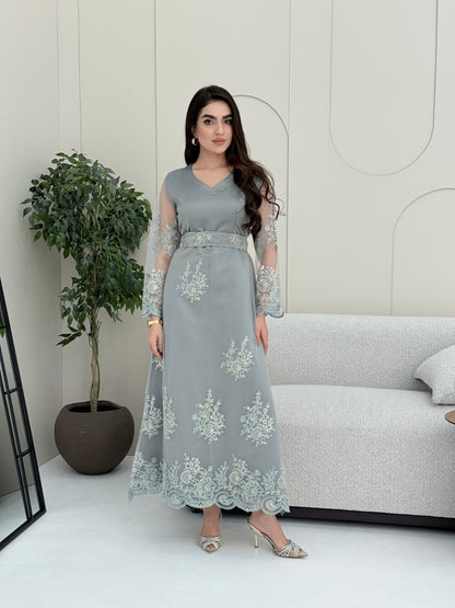 Woman wearing a light grey luxury jalabiya featuring delicate floral lace sheer sleeves, a tailored V-neck, and an elegantly embroidered matching waist belt, standing against a white wall with a plant in the foreground.