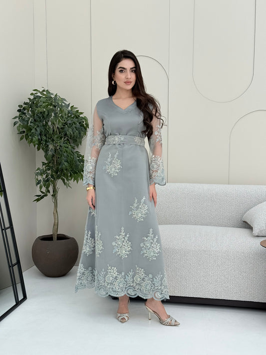 Woman wearing a light grey luxury jalabiya featuring delicate floral lace sheer sleeves, a tailored V-neck, and an elegantly embroidered matching waist belt, standing against a white wall with a plant in the foreground.