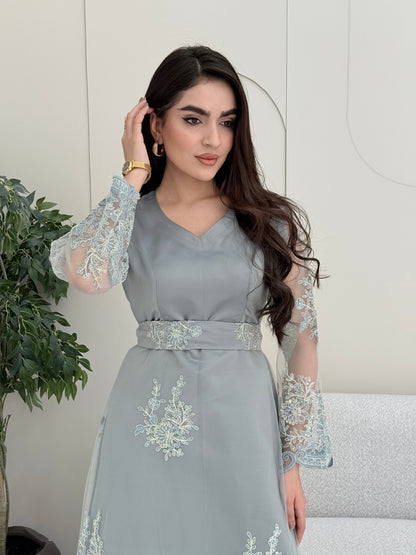 Woman wearing a light grey luxury jalabiya featuring delicate floral lace sheer sleeves, a tailored V-neck, and an elegantly embroidered matching waist belt, standing against a white wall with a plant in the foreground.