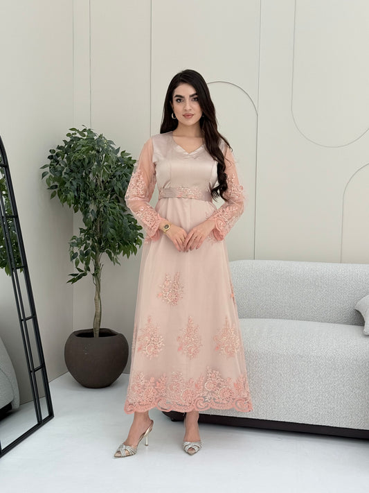 Woman in a beautiful light pink luxury jalabiya featuring shimmering floral embroidery on sheer sleeves, a flattering V-neck, and an intricately embroidered waist belt standing in a room with a plant and furniture.