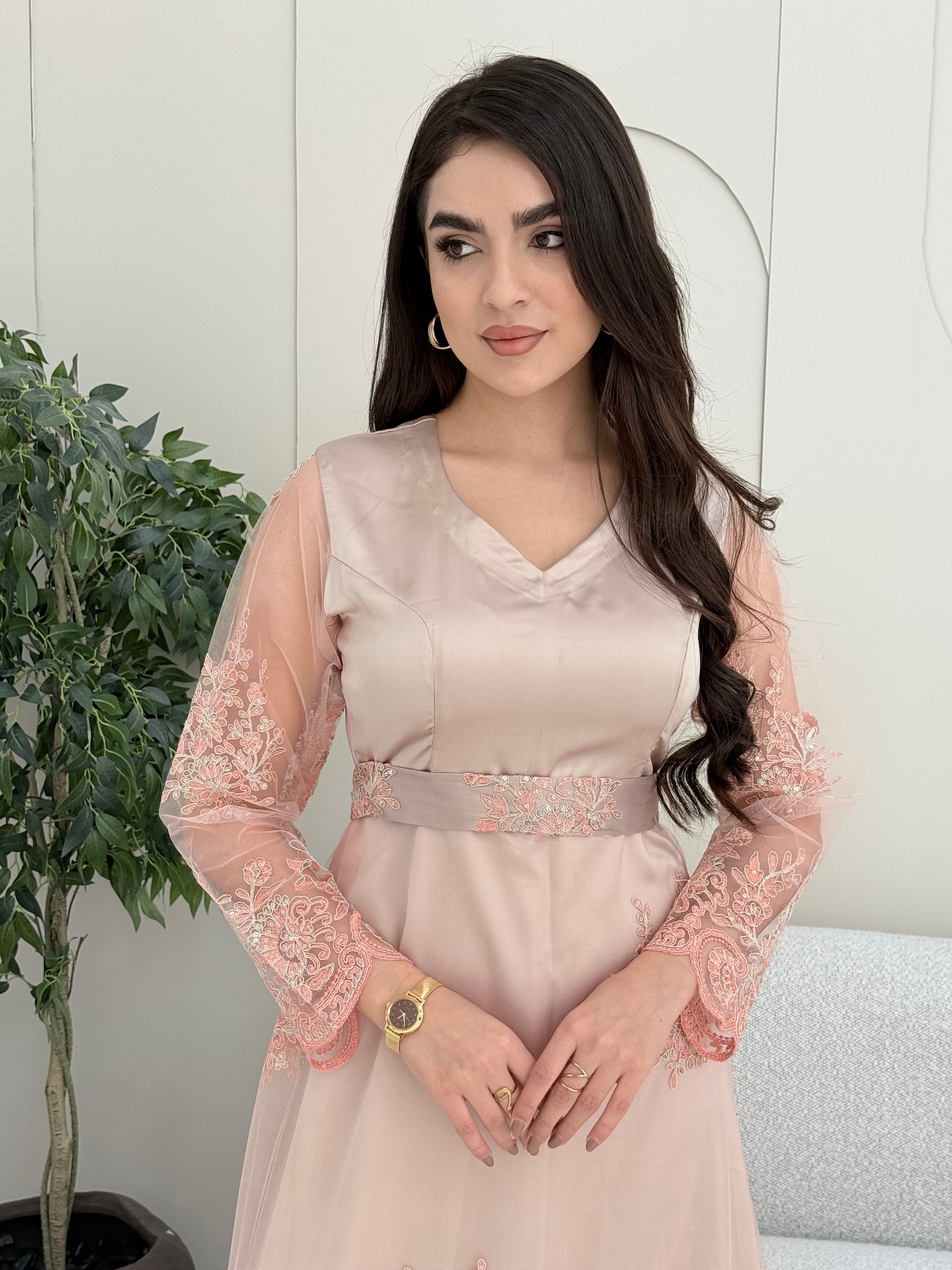 Woman wearing a beautiful light pink luxury jalabiya featuring shimmering floral embroidery on sheer sleeves, a flattering V-neck, and an intricately embroidered waist belt. against a white wall.