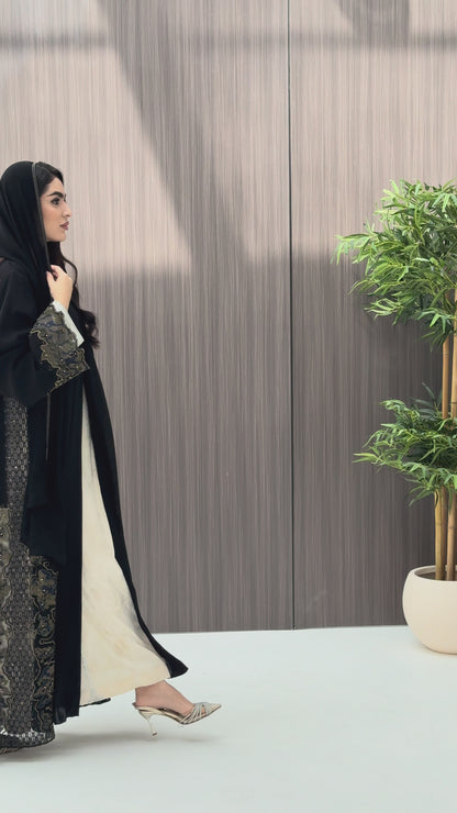 a woman wearing an elegant open-front black abaya featuring intricate multi-colored beadwork, sequins, and metallic embroidery in an indoor setting.